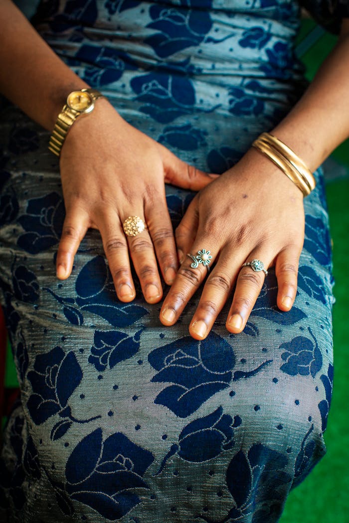 gallery-4 Close-up of hands adorned with rings and bracelets resting on a floral blue dress.