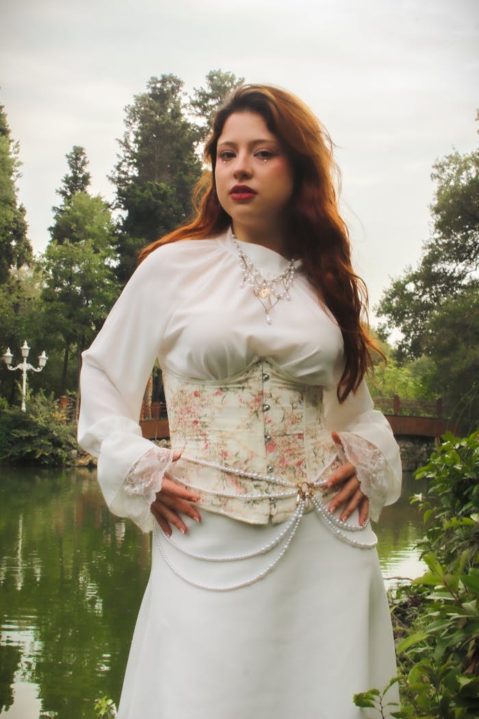 services-02 Woman in victorian-style attire posing gracefully by a serene lake in a lush garden setting.