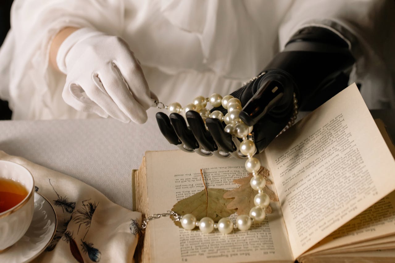 services-01 A graceful prosthetic hand holds a pearl necklace over an open book, blending technology and elegance.